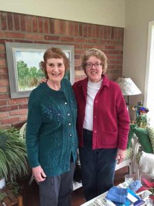 Rosemary Low standing with Susan Orosz in a room with a brick wall, painting and furniture